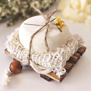 May include: A round, off-white soap bar sits on a wooden soap dish with a woven net and a wooden bead. The soap is tied with twine and adorned with a small yellow flower. The soap dish is made of small wooden slats.