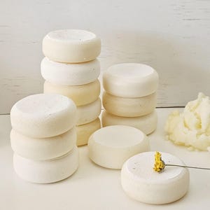 May include: Stacks of small, round, off-white objects, possibly soap or bath bombs, are arranged on a white surface. Some are stacked, while others are placed individually. A small yellow flower rests on one.