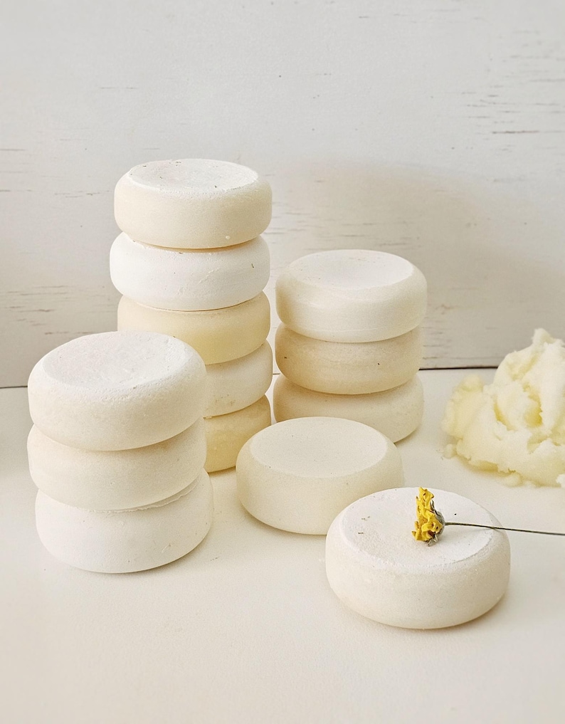 May include: Stacks of small, round, off-white objects, possibly soap or bath bombs, are arranged on a white surface. Some are stacked, while others are placed individually. A small yellow flower rests on one.