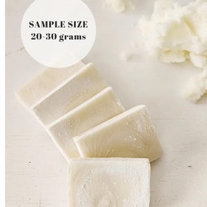 May include: A close-up of several rectangular bars of soap, arranged diagonally on a white surface. The soap is a creamy white color with a subtle marbled texture. The text "SAMPLE SIZE 20-30 grams" is in a white circle.