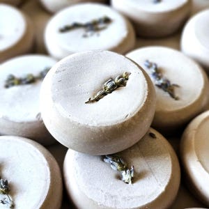 May include: Close-up of several round, light-colored bath bombs, each with a sprig of lavender. The bath bombs have a textured surface and are arranged in a slightly blurred, close-up shot, highlighting the natural elements.
