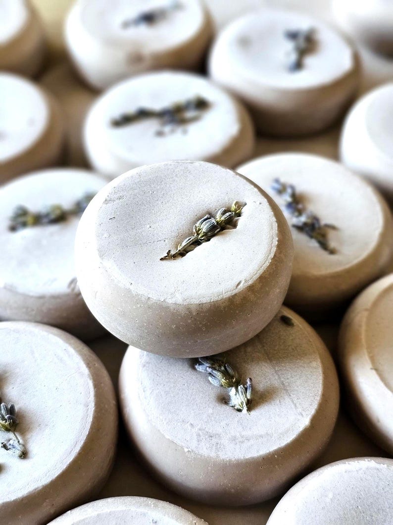 May include: Close-up of several round, light-colored bath bombs, each with a sprig of lavender. The bath bombs have a textured surface and are arranged in a slightly blurred, close-up shot, highlighting the natural elements.