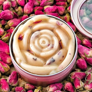 May include: A rose-shaped, beige-coloured soap bar with dried rose petals in a pink metal tin. The tin is surrounded by dried rose buds.
