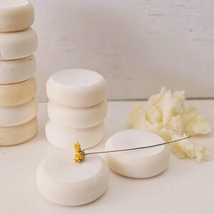 May include: Several stacks of small, round, off-white cheese wheels. One wheel has a small yellow flower attached with a thin wire. A pile of shredded cheese is to the right. The background is a white wooden surface.