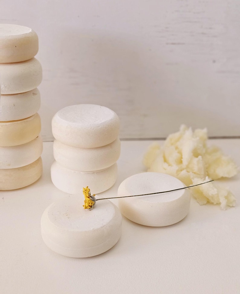 May include: Several stacks of small, round, off-white cheese wheels. One wheel has a small yellow flower attached with a thin wire. A pile of shredded cheese is to the right. The background is a white wooden surface.