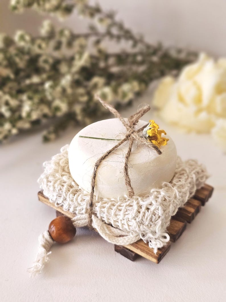 May include: A round, off-white soap bar sits on a wooden soap dish with a woven net and a wooden bead. The soap is tied with twine and adorned with a small yellow flower. The soap dish is made of small wooden slats.