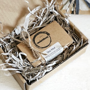 May include: A brown cardboard box with brown paper filler inside. The box contains a small brown cardboard box with a label that reads "Sea Mist" and lists the ingredients for the soap. The label also includes the company logo, "rawbatchscap".