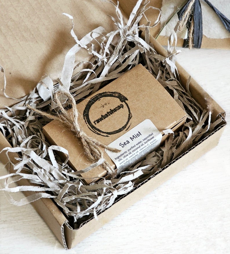 May include: A brown cardboard box with brown paper filler inside. The box contains a small brown cardboard box with a label that reads "Sea Mist" and lists the ingredients for the soap. The label also includes the company logo, "rawbatchscap".