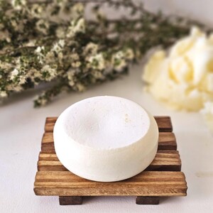 May include: A round, white bar of soap rests on a small, wooden soap dish. The dish is made of several parallel wooden slats. The background includes blurred white flowers and greenery.