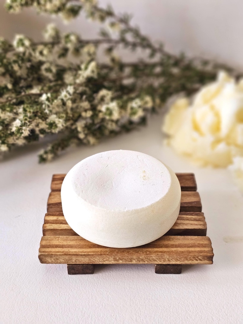 May include: A round, white bar of soap rests on a small, wooden soap dish. The dish is made of several parallel wooden slats. The background includes blurred white flowers and greenery.