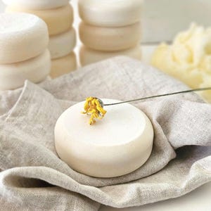 May include: Stacks of round, off-white soaps are arranged on a neutral-toned linen cloth. A single soap is adorned with a small yellow flower. The soaps have a smooth, clean appearance, suggesting a natural skincare product.
