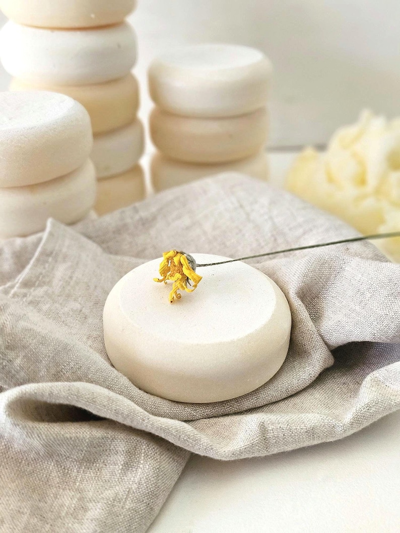 May include: Stacks of round, off-white soaps are arranged on a neutral-toned linen cloth. A single soap is adorned with a small yellow flower. The soaps have a smooth, clean appearance, suggesting a natural skincare product.