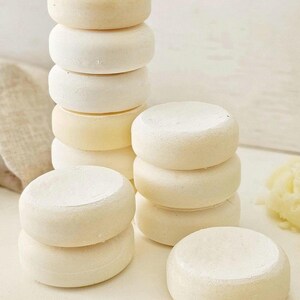 May include: Several stacks of small, round, off-white cheese wheels. The cheese wheels are stacked in towers of varying heights, with some individual wheels scattered around. The cheese has a slightly textured surface and a pale yellow hue.