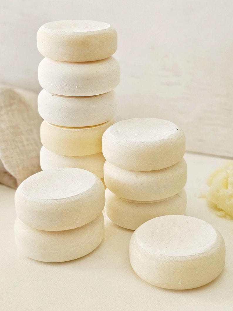 May include: Several stacks of small, round, off-white cheese wheels. The cheese wheels are stacked in towers of varying heights, with some individual wheels scattered around. The cheese has a slightly textured surface and a pale yellow hue.