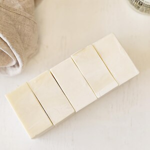 May include: Six rectangular bars of light-colored soap arranged diagonally on a white surface. A beige linen cloth is partially visible in the upper left corner. A jar with a black lid is in the upper right corner.