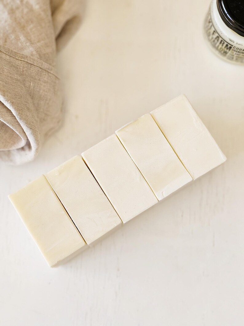 May include: Six rectangular bars of light-colored soap arranged diagonally on a white surface. A beige linen cloth is partially visible in the upper left corner. A jar with a black lid is in the upper right corner.
