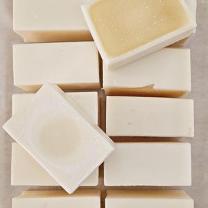 May include: An overhead shot shows several rectangular bars of soap. The bars are mostly white with a light yellow center. Some bars are stacked, while others are arranged in a grid pattern. The soap appears to be handmade.