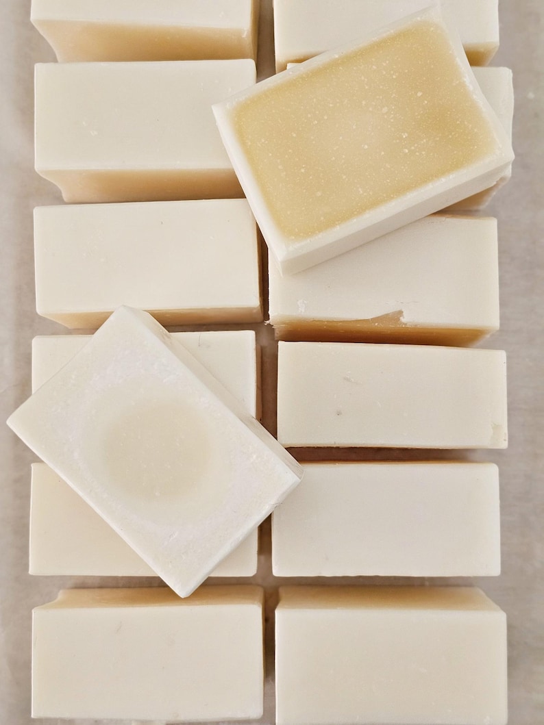 May include: An overhead shot shows several rectangular bars of soap. The bars are mostly white with a light yellow center. Some bars are stacked, while others are arranged in a grid pattern. The soap appears to be handmade.