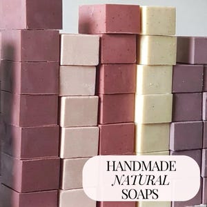 May include: A stack of handmade natural soaps in three colours: purple, pink, and white. The soaps are stacked in rows of four, with the text "HANDMADE NATURAL SOAPS" at the bottom of the image.