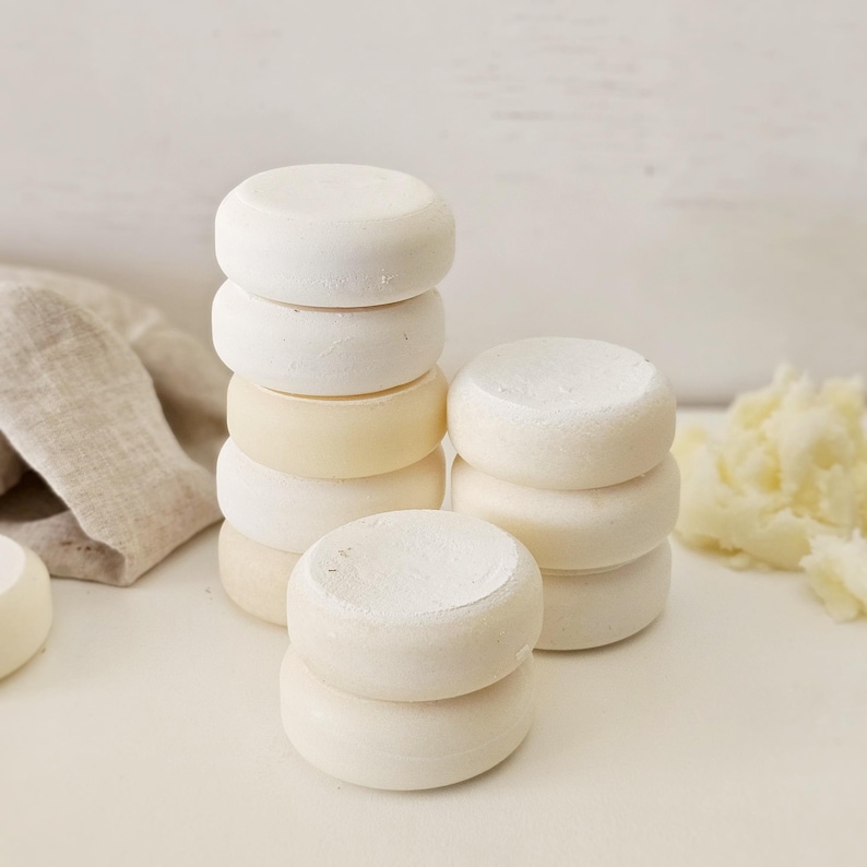 May include: Several stacks of small, round, off-white objects, possibly soaps or bath bombs, are arranged on a white surface. Some stacks are taller than others, and a few are a lighter shade of yellow. A linen cloth is visible in the background.