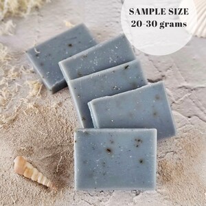 May include: Five bars of blue soap with flecks of grey, arranged in a stack on a light grey textured surface. The text "SAMPLE SIZE 20-30 grams" is visible in a white circle above the soap.