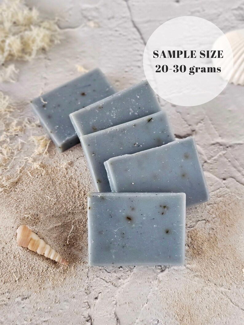 May include: Five bars of blue soap with flecks of grey, arranged in a stack on a light grey textured surface. The text "SAMPLE SIZE 20-30 grams" is visible in a white circle above the soap.