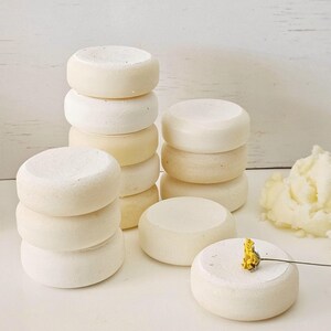 May include: Several stacks of small, round, off-white objects, possibly soaps or bath bombs, are arranged on a white surface. A small yellow flower rests on one of the objects. The items have a smooth, slightly textured appearance.