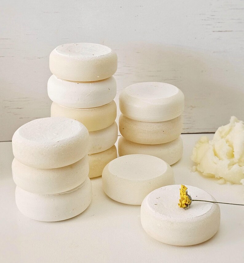 May include: Several stacks of small, round, off-white objects, possibly soaps or bath bombs, are arranged on a white surface. A small yellow flower rests on one of the objects. The items have a smooth, slightly textured appearance.