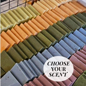 Op de afbeelding: Een assortiment handgemaakte zeepjes in verschillende kleuren, gestapeld in rijen. De zeepjes zijn gerangschikt in een rasterpatroon en hebben het label "Kies uw geur".