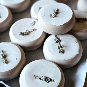 May include: Close-up of several round, light-colored bath bombs, each topped with a sprig of dried lavender. The bath bombs are arranged on a light-colored surface, creating a spa-like aesthetic. The lavender adds a touch of natural beauty and fragrance.