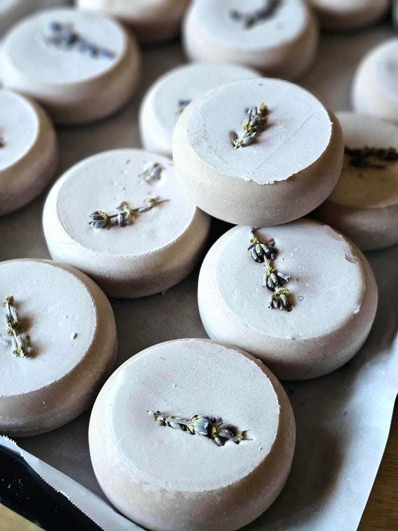 May include: Close-up of several round, light-colored bath bombs, each topped with a sprig of dried lavender. The bath bombs are arranged on a light-colored surface, creating a spa-like aesthetic. The lavender adds a touch of natural beauty and fragrance.