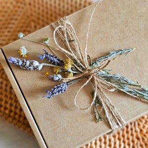 Puede incluir: Una caja de cartón marrón atada con cuerda y decorada con flores secas. Las flores son una mezcla de lavanda, amarillo y verde.
