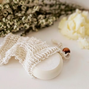 May include: A round, white bar of soap partially encased in a cream-colored, open-weave soap saver bag. The bag has a drawstring closure with a small, brown wooden bead. The background includes dried flowers and a soft, blurred texture.