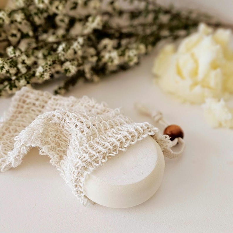 May include: A round, white bar of soap partially encased in a cream-colored, open-weave soap saver bag. The bag has a drawstring closure with a small, brown wooden bead. The background includes dried flowers and a soft, blurred texture.