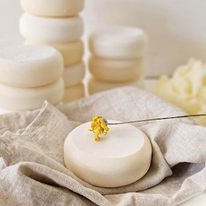 May include: Stacks of round, off-white soap bars are arranged on a neutral linen cloth. A single soap bar is adorned with a small yellow flower. The soaps have a smooth, clean appearance, suggesting a natural skincare product.