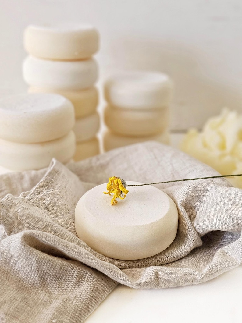 May include: Stacks of round, off-white soap bars are arranged on a neutral linen cloth. A single soap bar is adorned with a small yellow flower. The soaps have a smooth, clean appearance, suggesting a natural skincare product.
