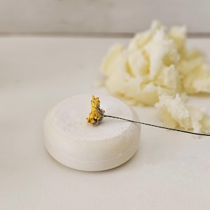 May include: A white, round soap bar with a small yellow flower on top. A pile of off-white, fluffy material is in the background. The soap bar is smooth and the flower has a delicate appearance.