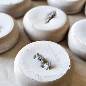 May include: Circular, light gray soap bars with a white top layer, each adorned with a sprig of lavender. The soaps are arranged on a light-colored surface, showcasing a natural, spa-like aesthetic.