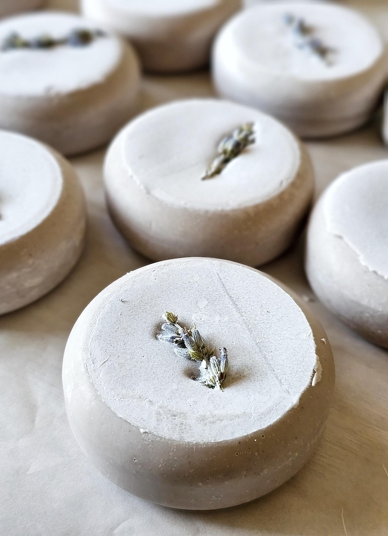 May include: Circular, light gray soap bars with a white top layer, each adorned with a sprig of lavender. The soaps are arranged on a light-colored surface, showcasing a natural, spa-like aesthetic.