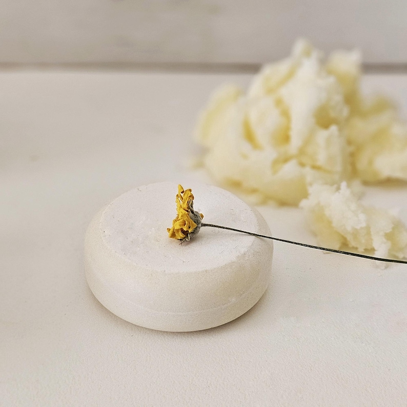 May include: A white, round soap bar with a small yellow flower on top. A pile of off-white, fluffy material is in the background. The soap bar is smooth and the flower has a thin, dark stem.