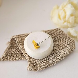 May include: A small, round, off-white ceramic soap dish with a dried yellow flower. The dish sits on a beige, woven washcloth. A blurred, light yellow, fluffy object is in the background.