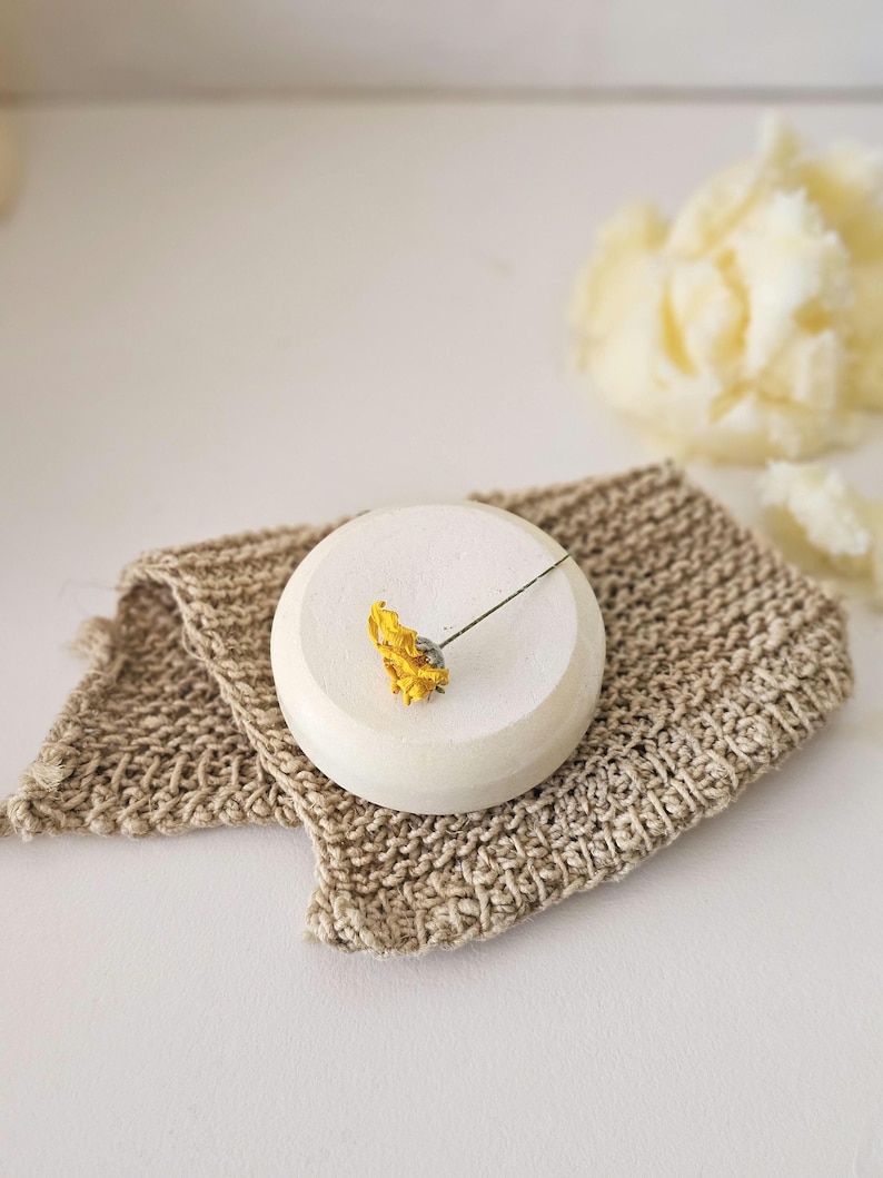 May include: A small, round, off-white ceramic soap dish with a dried yellow flower. The dish sits on a beige, woven washcloth. A blurred, light yellow, fluffy object is in the background.