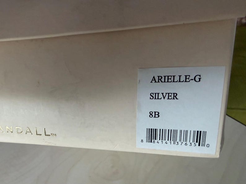 May include: A light beige rectangular cardboard shoe box with a white label on the side. The label features the text ARIELLE-G, SILVER, and 8B, along with a barcode. The partial text ANDALL is visible in gold lettering on the lower left side.