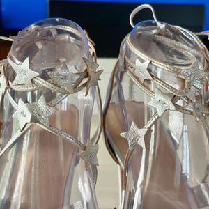 May include: A pair of metallic gold strappy sandals featuring multiple star-shaped cutouts along the ankle ties. The sandals are displayed on clear plastic forms, highlighting the intricate star detailing and the thin, wrap-around lacing design.