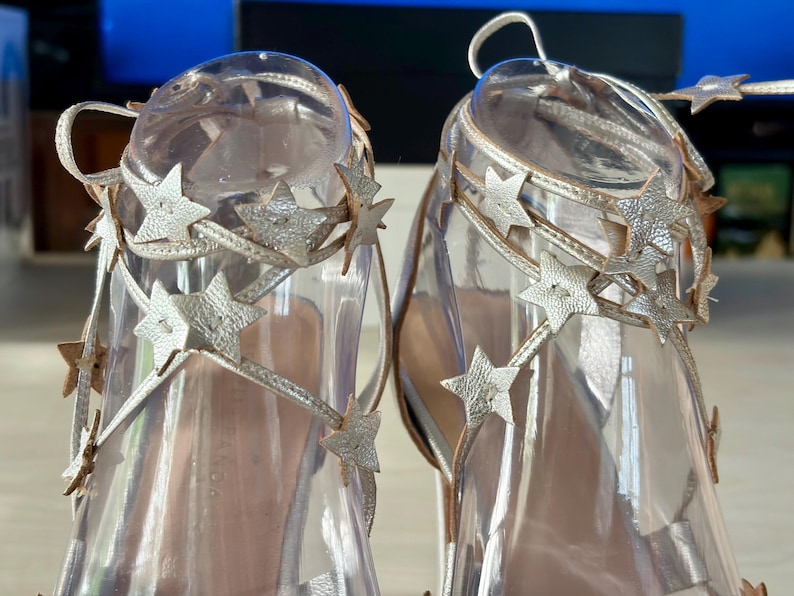 May include: A pair of metallic gold strappy sandals featuring multiple star-shaped cutouts along the ankle ties. The sandals are displayed on clear plastic forms, highlighting the intricate star detailing and the thin, wrap-around lacing design.