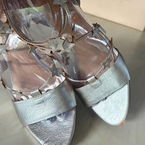 May include: A pair of metallic silver sandals featuring clear vinyl straps adorned with silver star-shaped embellishments. The footwear includes a wide toe band and ankle straps with a buckle closure, showcasing a textured finish on the leather.