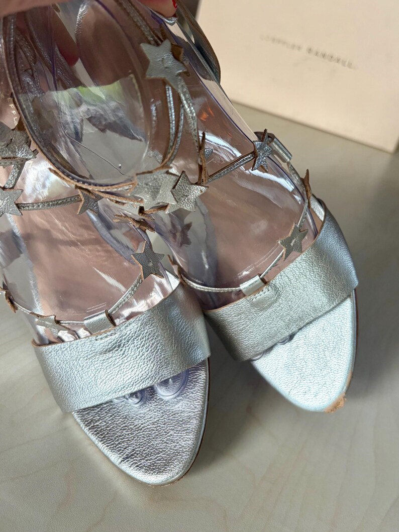 May include: A pair of metallic silver sandals featuring clear vinyl straps adorned with silver star-shaped embellishments. The footwear includes a wide toe band and ankle straps with a buckle closure, showcasing a textured finish on the leather.
