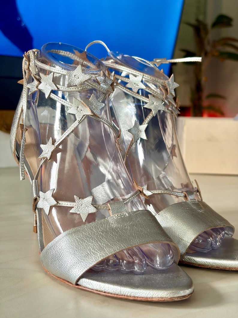 May include: A pair of metallic silver high-heeled sandals featuring a wide toe strap and thin, crisscrossing ankle straps adorned with multiple small, textured star-shaped cutouts. The shoes have a slim heel and a zip closure at the back.