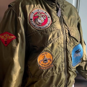 May include: An olive green military jacket featuring several patches. Patches include the US Marine Corps emblem, a red and gold emblem, and a patch that reads "Happy Intruders". The jacket has a zip closure and multiple pockets.