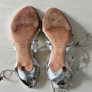 May include: A pair of silver metallic sandals with thin lace-up straps adorned with small star-shaped cutouts. The soles are tan leather with visible wear, featuring the Loeffler Randall brand name, a size 8 B stamp, and a small kitten heel.